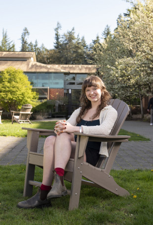 Bryn sitting in a chair outside with Watzek Library in the background. She is wearing a black dress with a white cardigan and brown boots.
