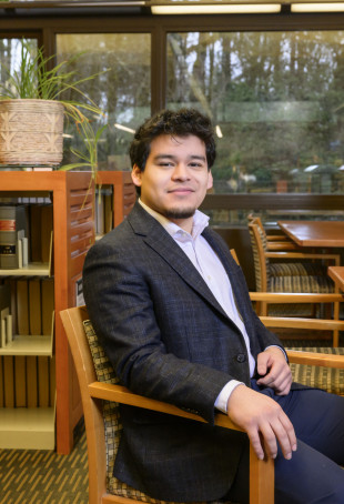 Manuel sitting on a chair in the library, wearing a dark grey blazer, white shirt, and jeans.