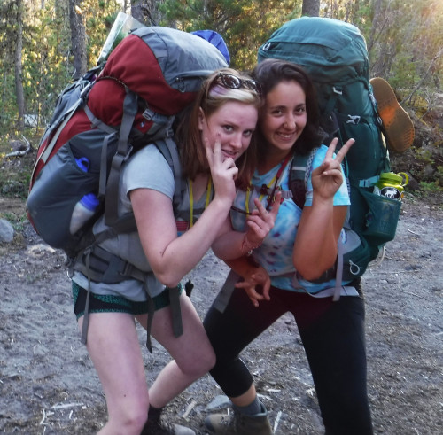 Image shows: two women wearing backpacking packs giving the peace sign and smiling.