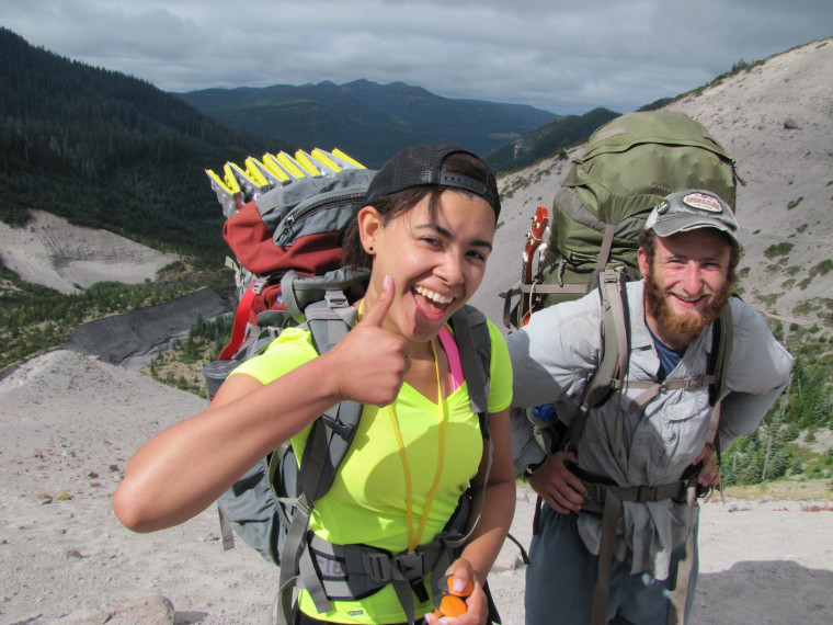 Image shows a person wearing a backpacking pack and a bright yellow shirt giving thumbs up to the camera. Another person stands to their ...