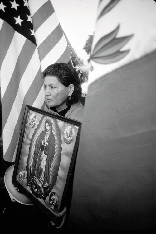 At a UFW rally, a woman holds an image of the Virgin of Guadalupe against a backdrop of the U.S. and Mexican flags.