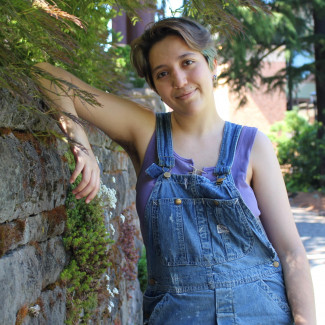 Fabi, in denim overalls, leaning against a stone wall.