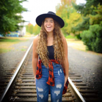 Ashtyn laughing and posing while standing on a train track, wearing a large black hat, black tank top, flannel shirt tied around waist.