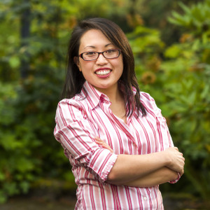 Sharon smiling outside, wearing glasses, a pink and white striped button-down shirt, and gray pants.