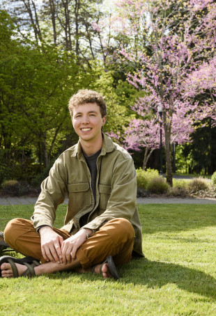 Max, sitting on the grass next to his backpack, smiling on a sunny day while wearing a green collared shirt, gray t-shirt and brown pants.