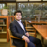 Manuel sitting on a chair in the library, wearing a dark grey blazer, white shirt, and jeans.