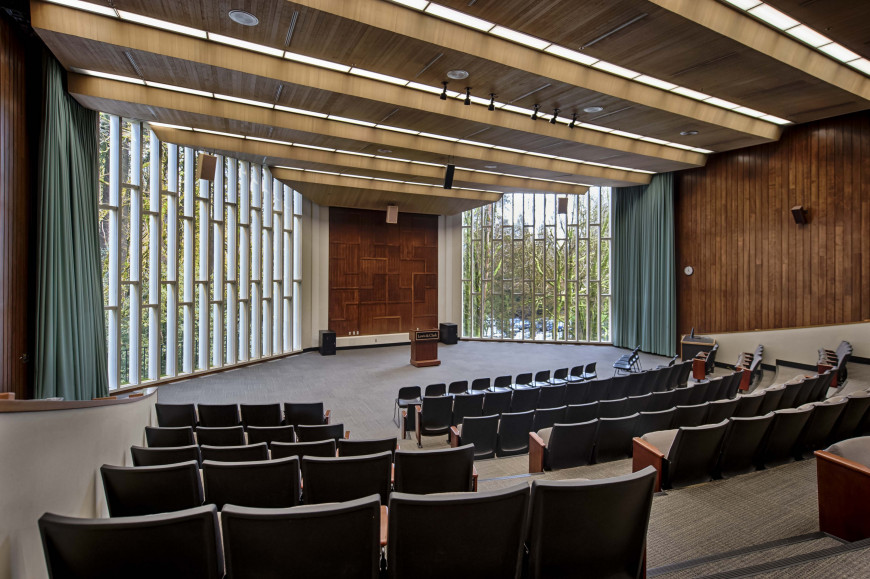 The open, tiered auditorium of the Council Chamber with wood back-wall paneling framed by large w...
