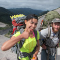 Image shows a person wearing a backpacking pack and a bright yellow shirt giving thumbs up to the camera. Another person stands to their ...