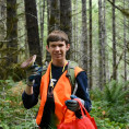 Person wearing bright orange vest and red shopping bag stands in the forrest. They are holding up large brown mushroom and smiling at the...