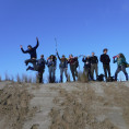Participants pose on a sand dune with their backpacks on and hiking poles in the air smiling with blue sky behind them.