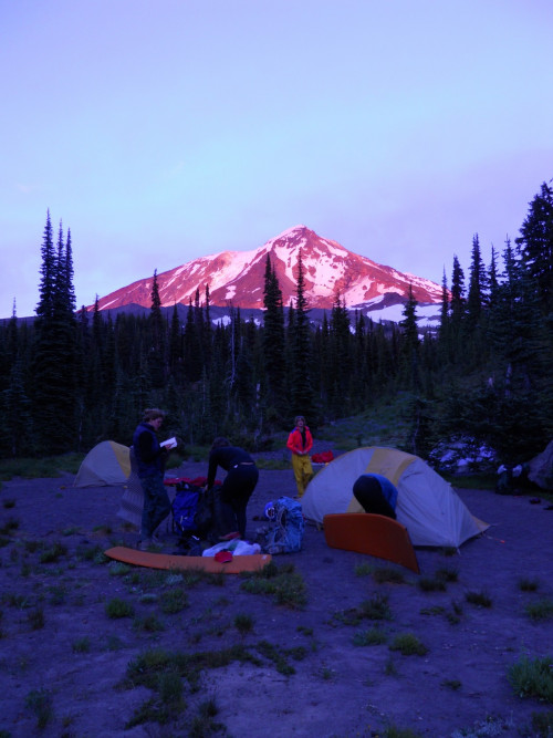 Image shows: a scene of tents and people milling around camp at sunset with Mt Adams lit up pink in the background.