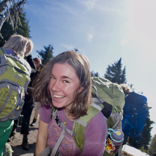 Image Description: Woman wearing a backpack smiles for the camera on a sunny day