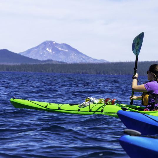 Image Description: Woman in a bright green kayak paddles on a blue lake with mountains in the bac...
