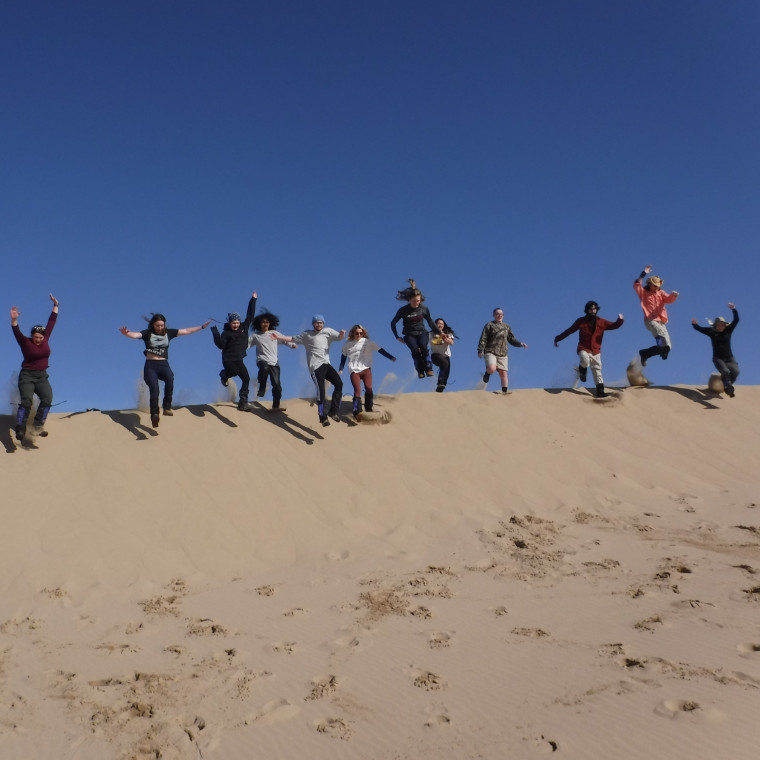Participants jump in the air with hands up on the sand dunes.