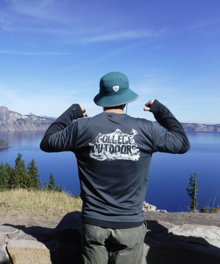 Person poses in front of Crater Lake with a teal sun hat and two thumbs pointing to the back of a grey College Outdoors shirt.