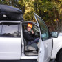 Image shows a man in an orange hat and black jacket sitting in a white vehicle. The door is open and he is giving a thumbs up and smiling.