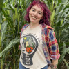 An image of Steph in a cornfield with her hands in her back pockets. She is where a white shirt t...