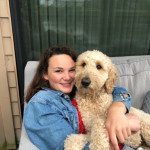 Christina is seated wearing a jean jacket and red shirt and hugging her goldendoodle.