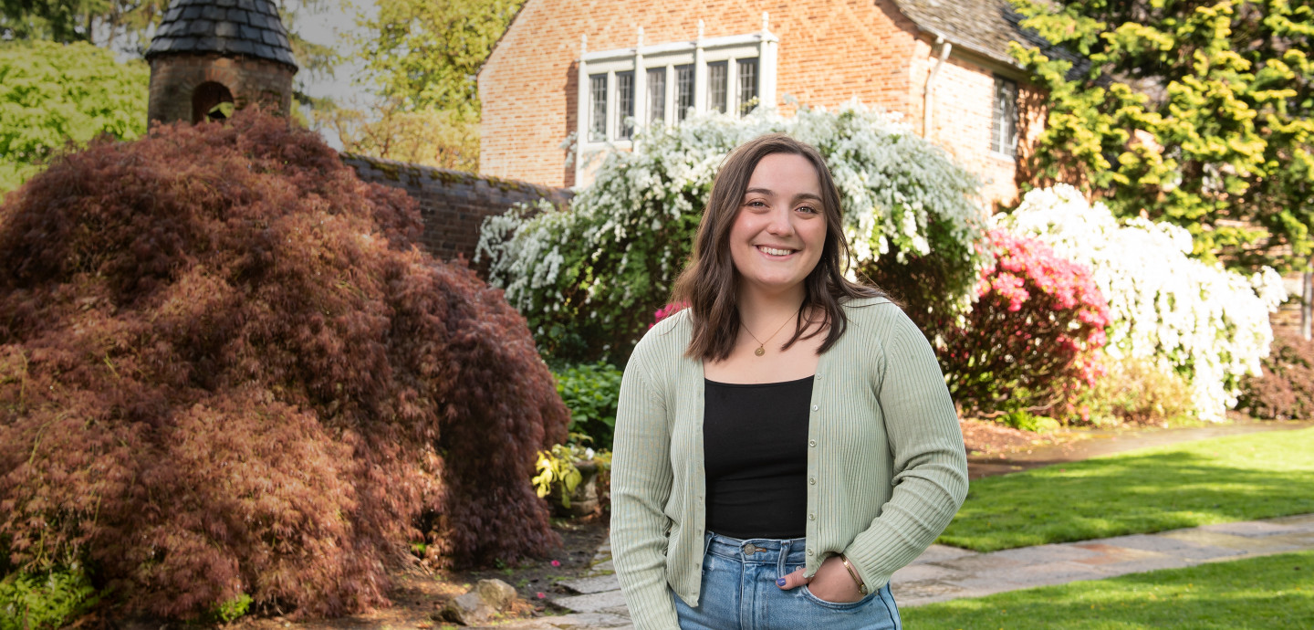 Greta Burton BA '22 poses outside the Manor House wearing a grey cardigan over a black top and blue jeans.