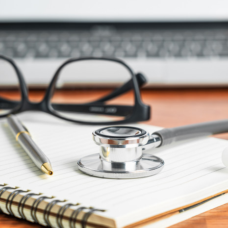 A journal with a medical stethoscope, pair of glasses, and pen on top, and laptop in the background.