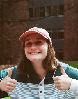 Alexandra Flory wearing a pink baseball hat, long-sleeved collared shirt, and giving two thumbs up to the camera while smiling.