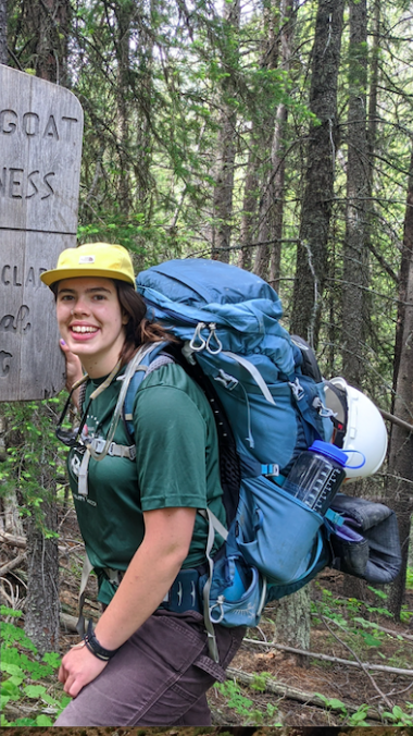Emily, smiling, standing at a hiking trail in the forest with a large backpack and walking stick.