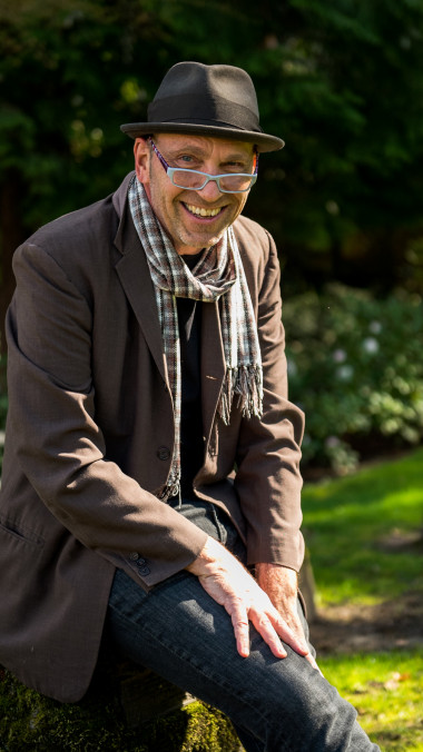 Štĕpán Šimek sits outside smiling and wearing a brown brimmed hat, glasses, a checkered scarf, and a brown jacket.