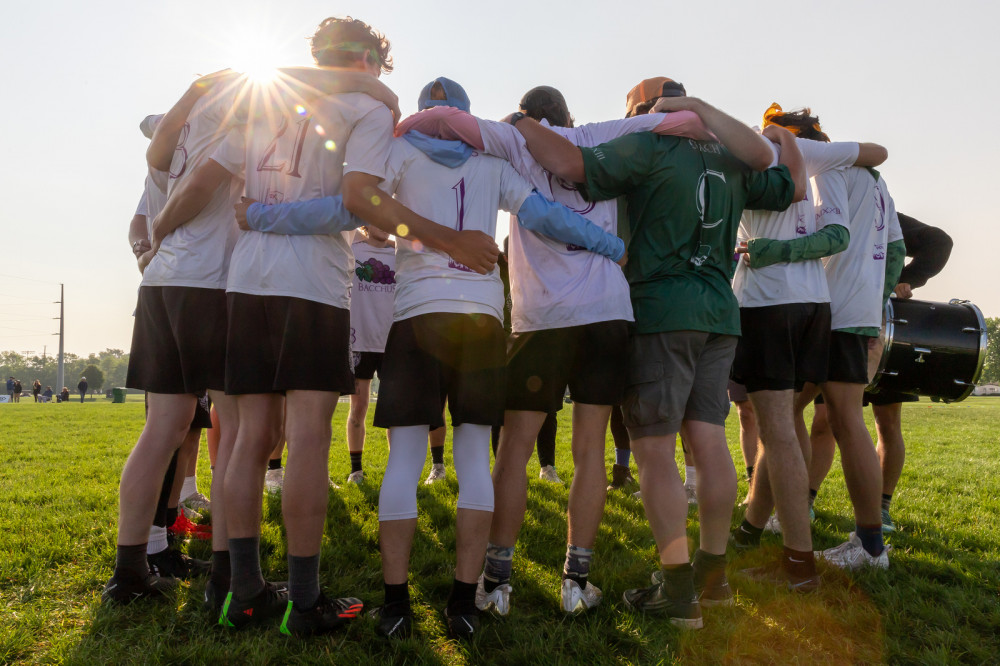 Backs of team members standing in a huddle with their arms around each other and sunlight shining above.