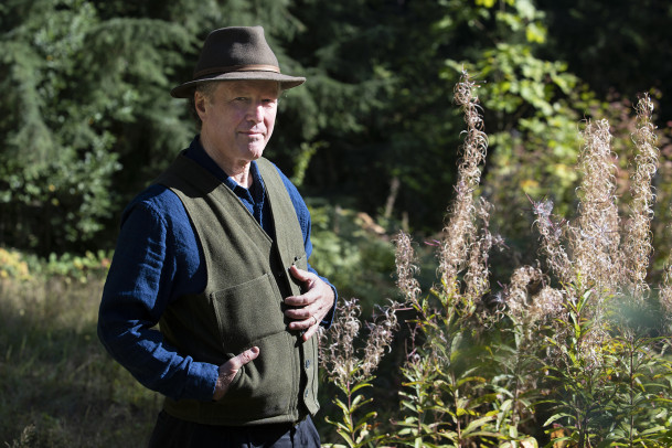 Kim Stafford posing outside, wearing a brimmed hat, green vest, and long sleeved blue shirt.
